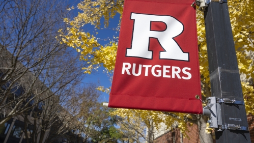 A red vertical banner featuring the Rutgers logo and branding flying during the day on a walkway at Rutgers Law School campus.