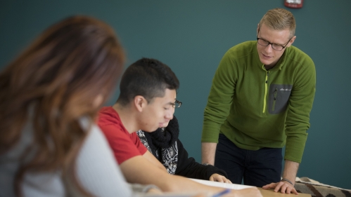 A spanish teacher closely instructs seated students in a classroom.