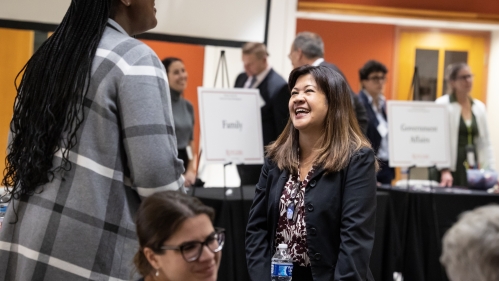 Two women engage over a table at a professional event in a large, busy balroom.