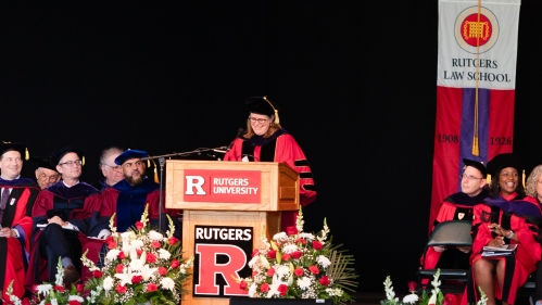 people in red commencement regalia seated on stage with woman at podium