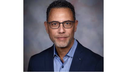 man in dark blazer and light blue shirt wearing glasses smiling against photo backdrop