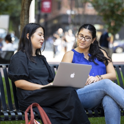 Two female students on bench outside law school looking at laptop