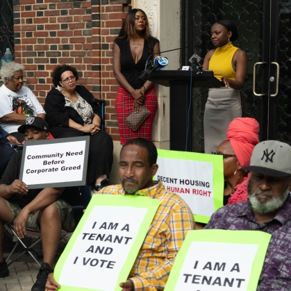 People sitting in folding chairs with signs at press conference