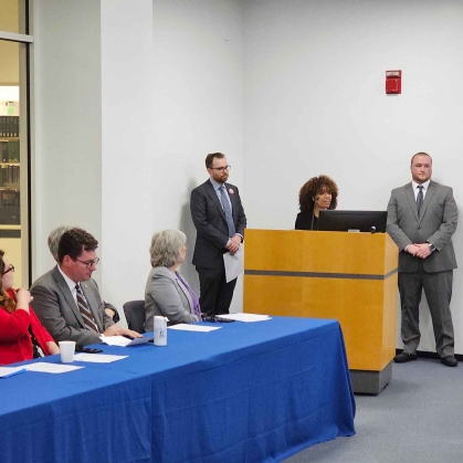 Student at lectern flanked by two men with people seated and listening at long blue table.