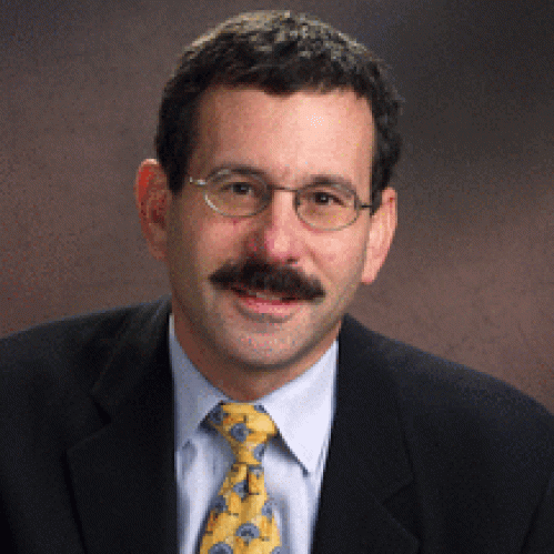 Man grinning wearing glasses, blue shirt, yellow tie, and suit jacket