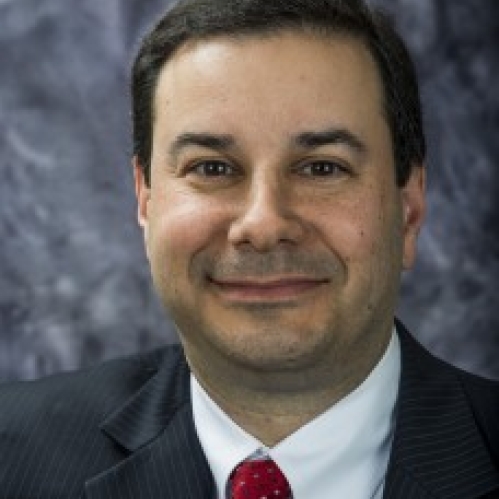 Man in dark suit, red tie, and white shirt smiling in front of grayish photo backdrop