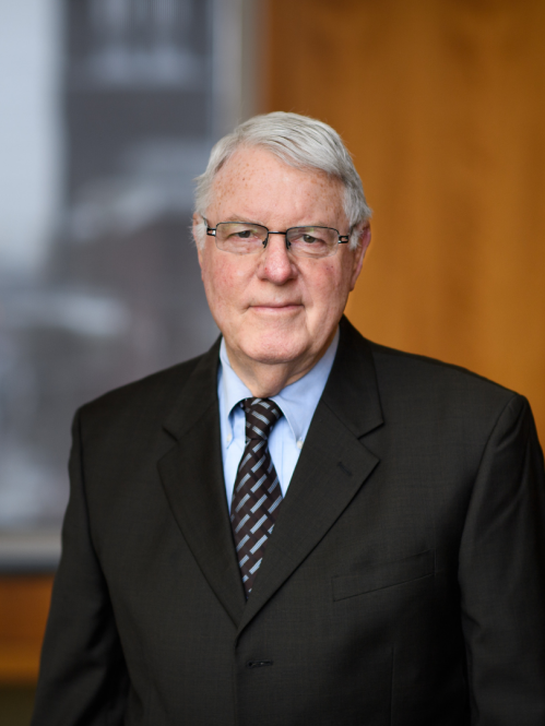 Man grinning wearing glasses, a blue button down shirt, a grey and black tie and a suit.
