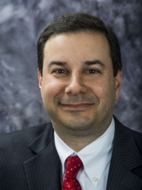 Man in dark suit, red tie, and white shirt smiling in front of grayish photo backdrop