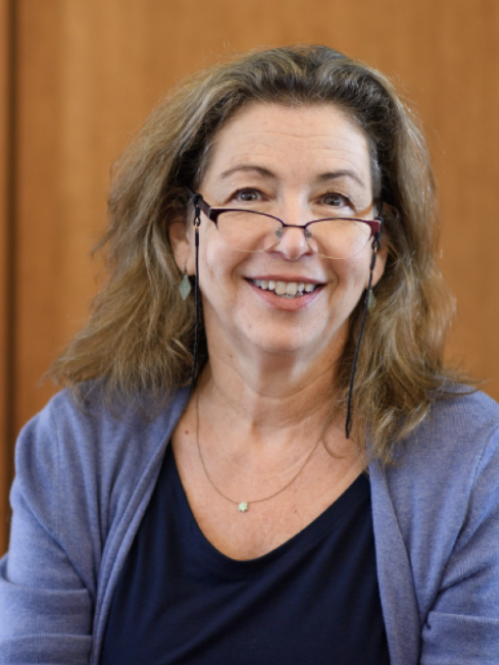 Woman smiling wearing glasses, a dark purple shirt, and a lilac cardigan. 