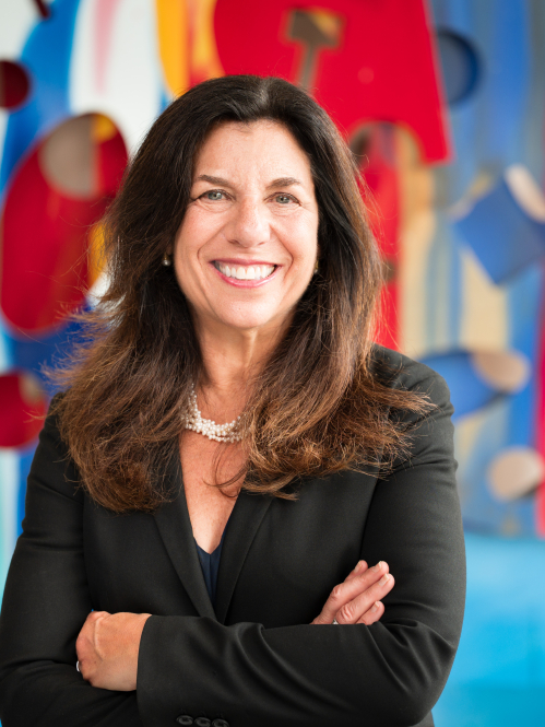 woman in black blazer smiling in front of colorful art with arms crossed