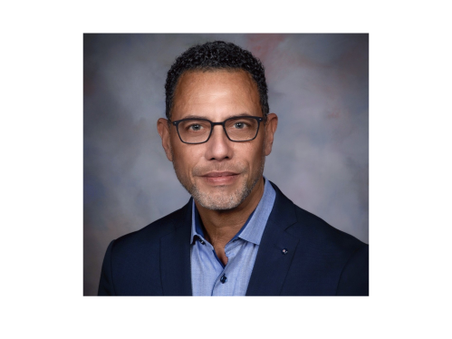 man in dark blazer and light blue shirt wearing glasses smiling against photo backdrop