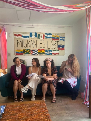 four women seating on a couch under 'migrantes LGBT' sign with various flags