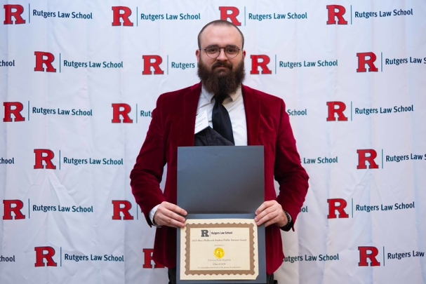 man wearing glasses and beard holding certificate and smiling in front of Rutgers Law School background