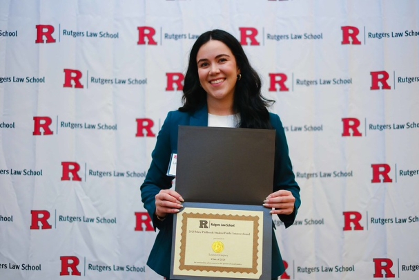 woman with long hair holding certificate and smiling in front of Rutgers Law School background