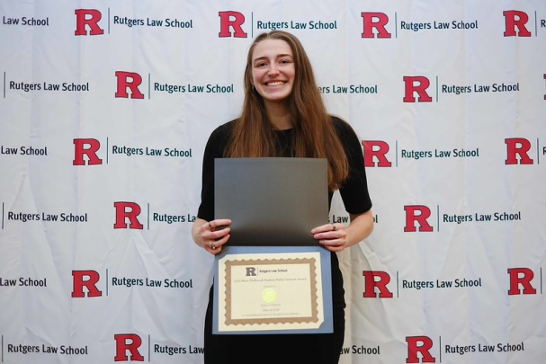 woman with long hair holding certificate and smiling in front of Rutgers Law School background