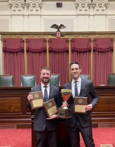 Two men in business suits smiling and holding plaques and a trophy in courtroom