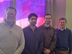 four men standing side by side and smiling in front of colorful window