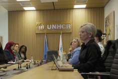 Students seated at a long conference table listening during a meeting, with a UNHCR sign and UN flags on the wall behind them.